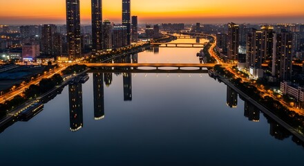 Fototapeta premium Aerial view of cityscape at dusk reflecting lights on water with buildings and bridges in the foreground under an orange sky