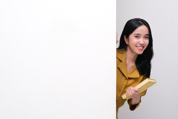 Indonesian female teacher peeking behind wall holding a book with happy face