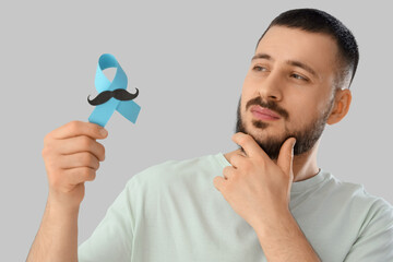 Thoughtful young man with blue ribbon and paper mustache on light background, closeup. Prostate...