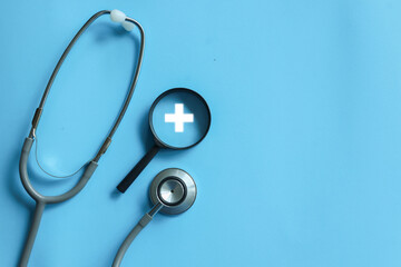 close up top view on magnifier with cross sign and stethoscope on blue background for health care checkup concept