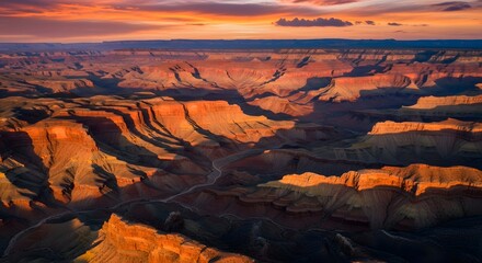 Aerial view of a vast canyon landscape bathed in warm sunlight creating dramatic shadows and highlighting the textured rock formations with vibrant orange and red hues under a sunset sky.
