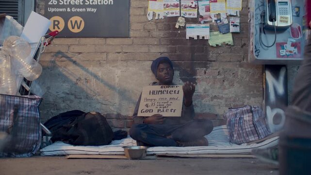 Young Black man sitting on street near subway, holding cardboard sign asking for money while people walking past him without noticing