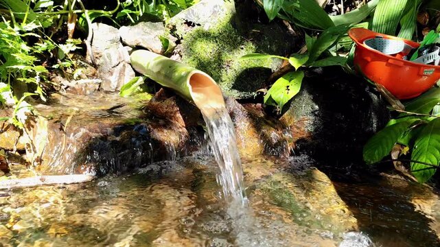 Clear spring water flows through a bamboo pipe into a rocky stream, surrounded by lush greenery and sunlight. A bright orange basin adds a touch of human presence to this peaceful nature scene.