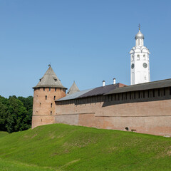 Brick defensive towers and the white-stone bell tower of the Novgorod Kremlin stand against a clear...