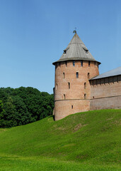 Historic round tower of the Novgorod Kremlin with wooden conical roof, surrounded by lush green landscape under a clear blue sky.