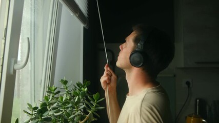 Young male listening to music, gently raising kitchen window blinds, welcoming natural sunlight and embracing peaceful morning moment with calm, contemplative demeanor - Powered by Adobe