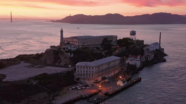 Alcatraz Island, USA, at dusk. The former prison, now a tourist destination, sits in the bay, with the lighthouse beacon shining. The sky is pink and purple.