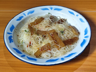A dish of fried vermicelli with pieces of kikil, sliced chilies and vegetables, served on a blue and white patterned enamel plate on a wooden table.