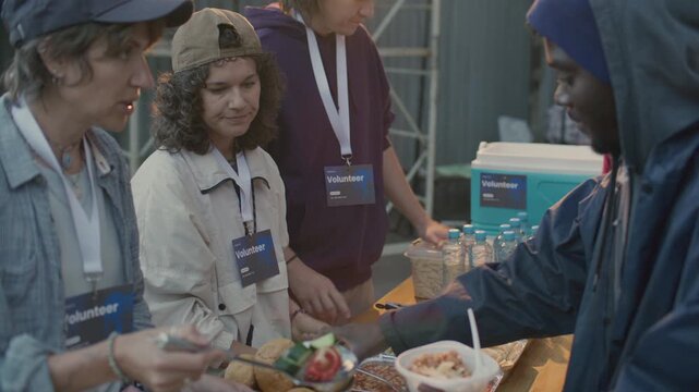 Group of volunteers wearing badges giving hot meals to hungry homeless people at food station during outreach program on urban street