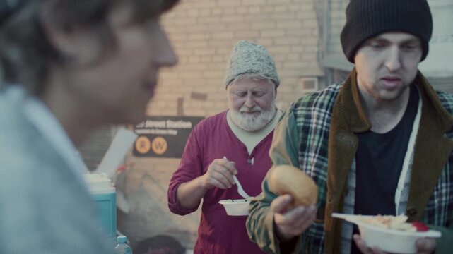 Diverse group of homeless men receiving hot meals and bread from volunteers and eating at food station in urban back alley