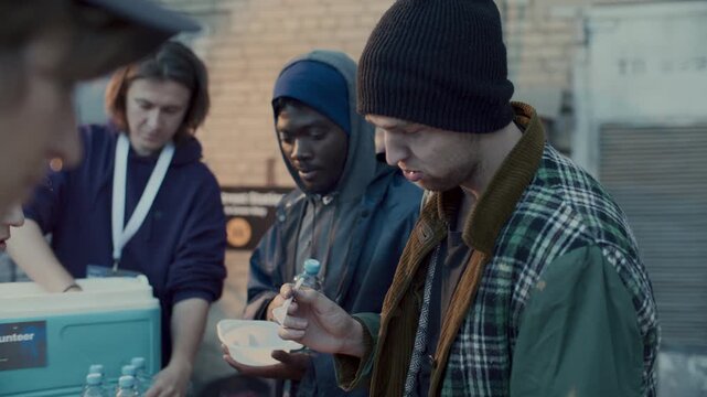 Multi-ethnic homeless people receiving water bottles and bowls of hot meal from volunteers at food drive during outdoor relief event in urban street