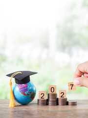 Earth globe with graduation hat, Wooden block with number 2027 on stack of coins. The concept of saving money for education, student loan, scholarship, tuition fees in year 2027.