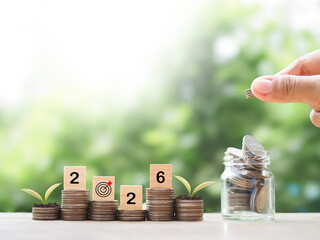 Hand putting coin into glass bottle, Wooden blocks with the number 2026 and plants growing up on stack of coins. The concept of saving money, Financial, Investment and Business growing in year 2026.