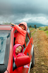 Two little Asian sisters peeking out from car window and smiling, enjoying their adventurous roadtrip  through the mountain track