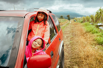 Two little Asian sisters peeking out from car window and smiling, enjoying their adventurous roadtrip  through the mountain track