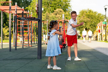 Boy in red shorts and younger girl in a blue dress chase floating bubbles near climbing bars at a...