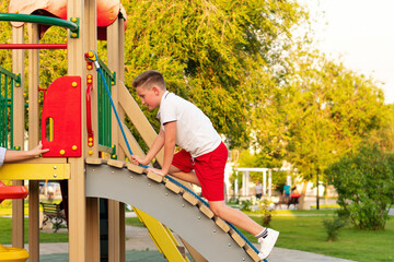 Determined boy in red shorts hauls himself up curved ramp with blue rope at sunny park playground,...
