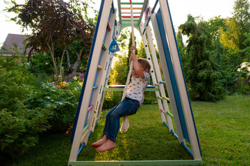 Barefoot boy hangs from thick white rope inside a colorful climbing frame, evening sun glowing...