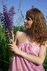 Woman in pink dress holding purple flowers outdoors amidst tall green grass