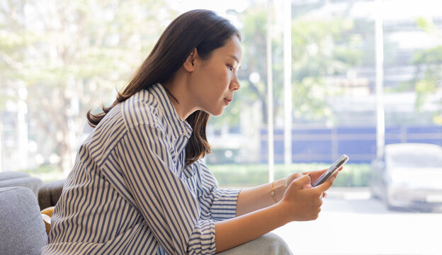 young asian businesswoman hold smartphone and looking on device screen for check notification or read news or alert from application in living room at home for people lifestyle and technology concept