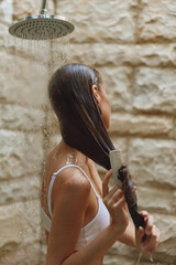 Woman washing long wet hair under shower with stone wall background, wearing white top, natural...