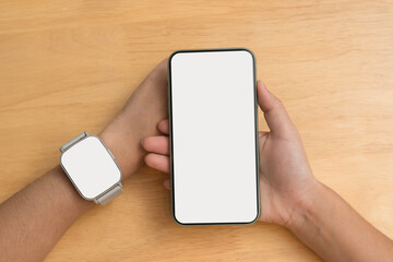 Hand holding smartphone with blank white screen and smart watch on wrist over wooden table background