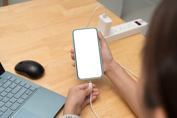 Person holding smartphone with blank screen and connecting charger while working on laptop at wooden desk