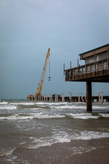 Vertical color image of a construction site on a pier along the Texas Gulf Coast. A large yellow crane stands at the end of the pier, framed by crashing ocean waves and overcast skies.