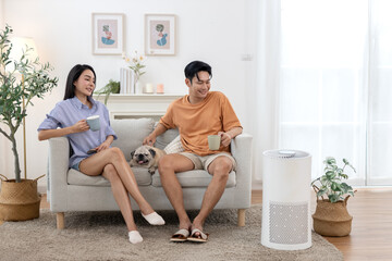 young asian couple drinking morning coffee sitting on sofa relaxing time living room with small pug dog rests beside them with air purifier for clean air in house for good health.