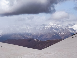 A landscape features snow-covered peaks with a dramatic sky filled with clouds. The rugged terrain...