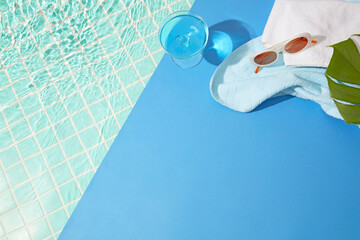 Calm poolside with blue tiles, white towel, sunglasses, and a cool blue drink, leaving a serene open space for creative summer displays.