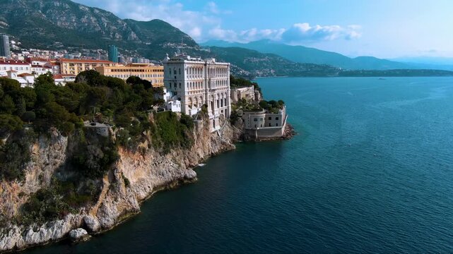 Dramatic drone footage of Monaco&rsquo;s famed Oceanographic Museum rising from sheer limestone cliffs above the turquoise Mediterranean, with Monte‑Carlo skyline and Alpine foothills beyond.