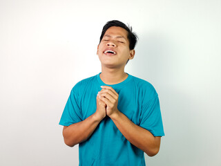 A young Asian man wearing a blue shirt smiles contentedly, clasping their hands together in...