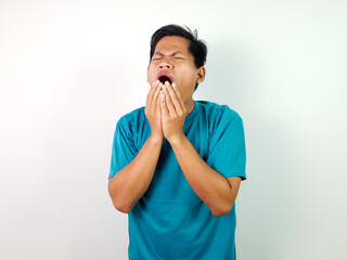 A young Asian man wearing a blue shirt yawns and covers his mouth with his hands, expressing...