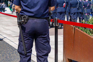 Police officer in uniform with baton and radio on the belt, standing by a red security tape during an official ceremony in Bydgoszcz, with uniformed officers in formation in the background.