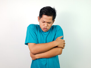 A young Asian man in a teal shirt hugging himself while seeming uncomfortable against a white...