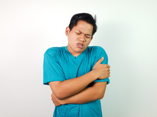 A young Asian man in a teal shirt hugging himself while seeming uncomfortable against a white...