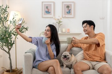 Happy young asian couple drinking morning coffee sitting on sofa relaxing time living room smiling use smartphone take selfies photo with small pug dog rests beside.