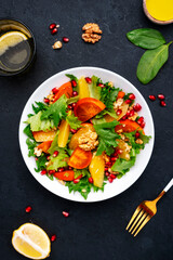 Autumn vegan persimmon salad with pomegranate seeds, walnuts, orange slices and lettuce, black background, top view