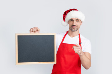 cheerful mature man in santa hat and apron hold blackboard with copy space. point finger
