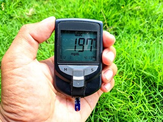 Close-up of a hand holding a digital glucometer showing 197 mg/dL blood sugar level, taken outdoors...