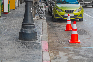 Urban street landscape with sidewalk: empty parking space on side of road with markings, place for car is marked with orange and red plastic traffic cones, there is a bicycle, a lamp post, yellow car