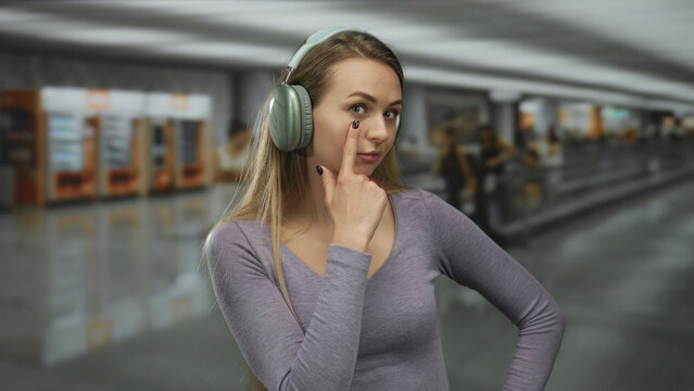Woman wearing headphones in airport making gesture with finger to indicate she is watching the surroundings, adding a playful element to the busy indoor travel atmosphere.