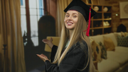 Woman smiling indoors wearing graduation cap in living room showcasing achievement with long blonde...
