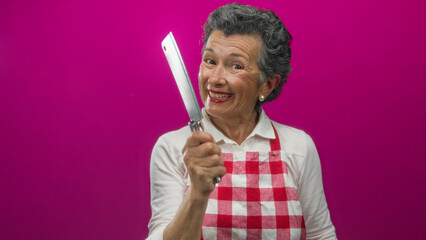 Senior woman in checkered apron smiling with knife in hand against vibrant pink background, exuding joyful cooking experience.