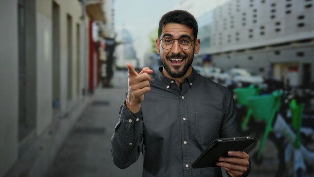 Smiling young man using a tablet and pointing confidently on an urban street, exuding charm and friendliness in an outdoor city environment.