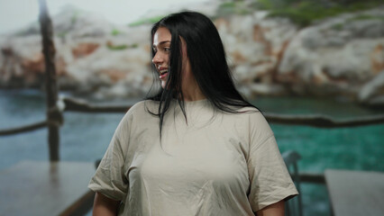 Young curvy woman smiling by the seaside with rocky cliffs in the background.