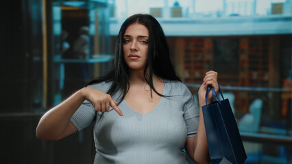 Young woman with serious expression points downward in a hotel lobby while holding a shopping bag,...
