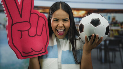 Woman smiling indoors holding soccer ball and foam glove in restaurant setting showcasing youthful...