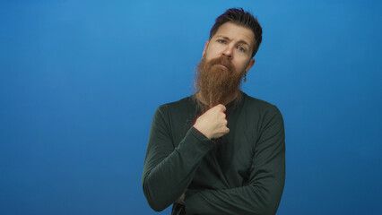Young caucasian man with long red beard and earrings crosses arms in blue studio; contemplation...
